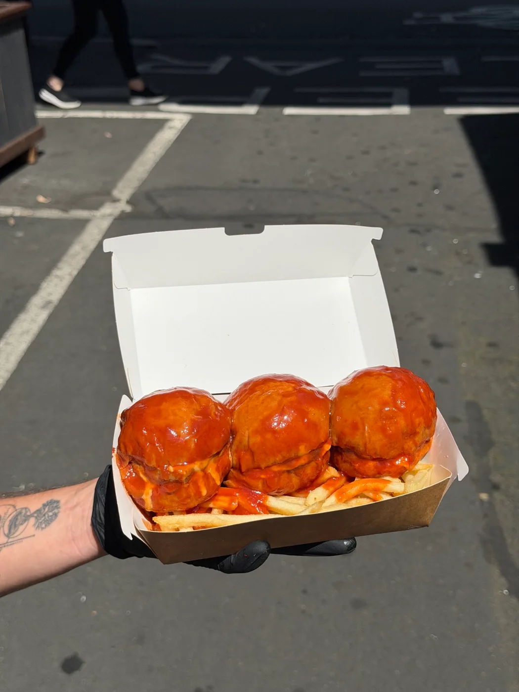 Three glossy tomato-sauced SOSMOS burgers and chips, held in a takeaway box